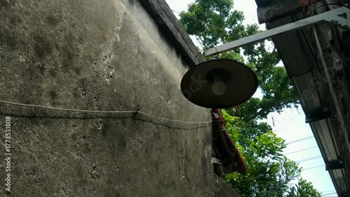 Footage of rustic outdoor lamp hangs on a weathered concrete wall, surrounded by trees and old rooftops under a bright daylight sky.