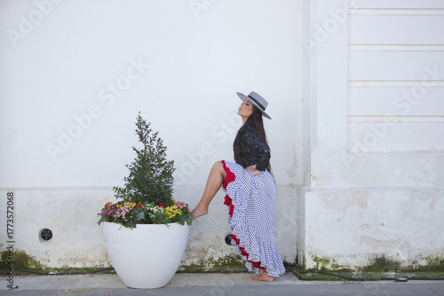 Young woman, beautiful, brunette, flamenco dancer, wearing a beautiful dress with jacket and hat, barefoot, posing with one foot on a flower pot on a white background wall.