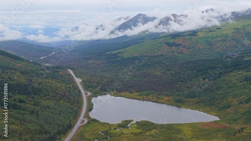 Aerial view of a winding road alongside a serene lake, surrounded by lush green forests and mountains shrouded in mist, Seward, Alaska, United States.