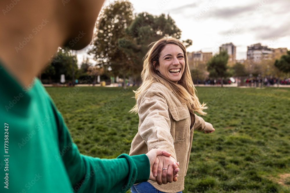 Obraz premium Woman smiling, looking back while holding hands with a person, pulling them forward in a green urban park