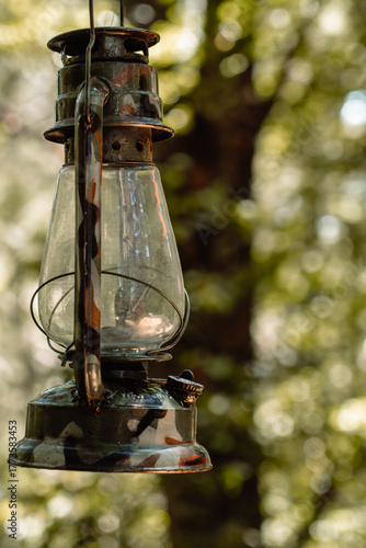 A vertical, close-up shot of a dark, rustic oil lantern hanging outdoors. The scene features a bright, soft-focus green and yellow background, highlighting the vintage object with warm, natural tones