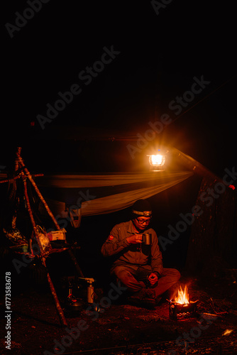 A man sits cross-legged, holding a mug and sipping a drink next to a small campfire and bright lantern at night. The image has a very dark, warm, high-contrast tone, showing cozy solitude