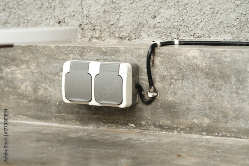Dual outdoor electrical outlet with protective covers installed on a concrete wall, featuring weatherproof casing and visible wiring conduit.
