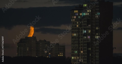 A time-lapse shot of an orange crescent moon setting over the night sky of Kyiv, Ukraine. Illuminated residential buildings contrast with the glowing lunar surface.