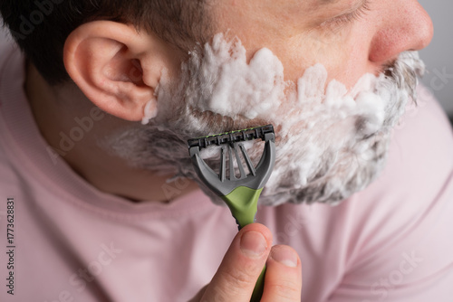Man shaves his beard with a razor and shaving foam. Morning routine and self-care at home. Men's grooming, clear skin, freshness, and confidence. Hygiene, beauty, and self-care concepts for men.
