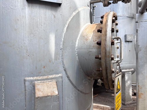 Close-up of a large industrial metal storage tank or vessel featuring a bolted flange connection. The heavy-duty steel component is secured with many rusty bolts and nuts, with a caution sign hanging 