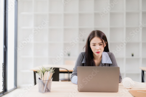 Asian businesswoman focusing intently on her laptop computer, working or studying at a wooden desk in a bright, modern office with a minimalist design and natural light