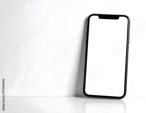 A black smartphone with a blank white screen leans against a white wall, reflecting on a glossy surface.