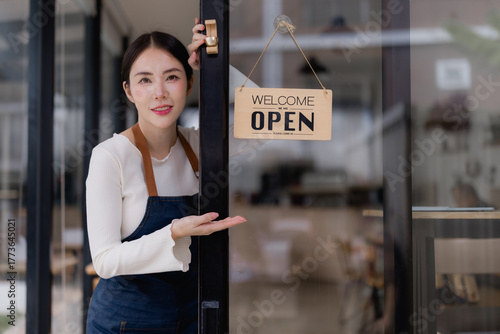 Smiling Asian woman small business owner wearing an apron, opening the glass door of her coffee shop and gesturing, displaying an open sign to welcome customers