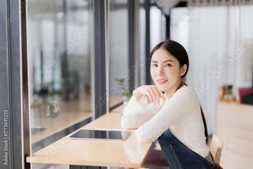 Young Asian woman small business owner smiling confidently, sitting at a cafe table with a digital tablet, representing entrepreneurship, modern business management and hospitality Free Download Young Asian woman small business owner smiling confidently, sitting at a cafe table with a digital tablet, representing entrepreneurship, modern business management and hospitality - GFXLoad