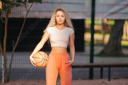 Confident young woman in sportswear holds a basketball on a fenced city court, soft evening light. Clean athletic portrait for fitness, sport and lifestyle uses.