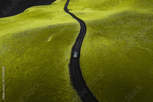 Aerial view of a lone car winding through a vibrant green landscape cut by a stark dark road under a brooding sky, Highlands Of Iceland, Sveitarfélagio Hornafjorour, Iceland.