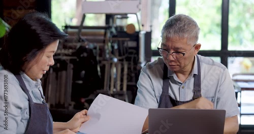 Asian elderly female and male cafe owners in aprons reviewing business accounting report together at cafe table, teamwork communication highlighting cooperation partnership and financial management
