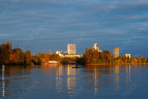 Beautiful autumn landscape at golden hour in Herastrau Park in Bucharest with boats on lake, colorful trees and office towers in the distance