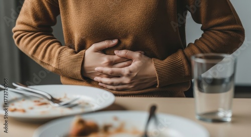 Person experiencing stomach ache after eating, holding their abdomen with hands, blurry table with dirty plates and glass of water