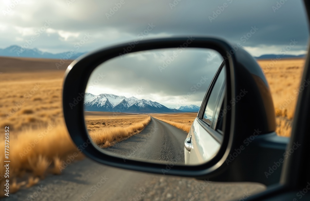Fototapeta premium Car side mirror view shows rural asphalt road leading to distant snow capped mountains and dramatic sky. Autumn field on road side. Trip in nature on scenic highway.