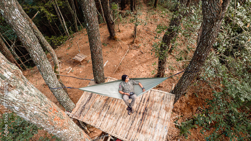 High-angle shot. A man lounges in a hammock tied between trees on a bamboo platform in a dense, sunlit forest. The scene features warm, earthy tones and a peaceful atmosphere