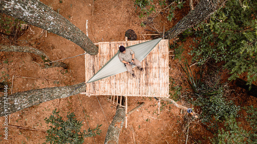 Aerial perspective. A man sits on a rustic bamboo platform built around four trees in a forest. The scene has an earthy, warm tone under soft natural light