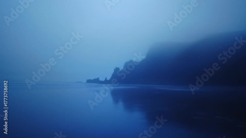 Ethereal blue coastal landscape with rugged cliffs shrouded in heavy mist, reflecting on tranquil ocean surface, mysterious and serene nature scene.