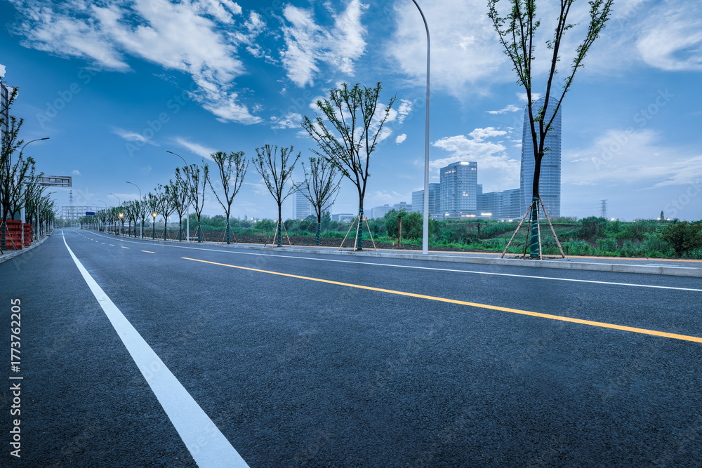 Naklejka premium Empty winding asphalt road through a modern urban park area with city buildings in the background under a blue sky.