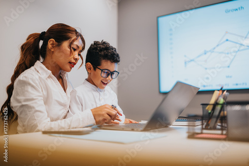A woman and a child work together at a modern office desk, reviewing notes and a laptop as a large screen shows a rising graph, conveying collaboration, learning, and business growth.