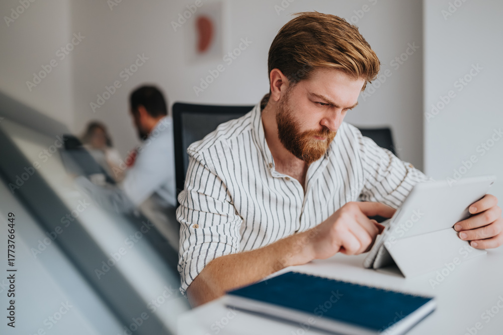 Naklejka premium A bearded man studies content on a tablet in a modern office. Background colleagues and a clean workspace convey a professional, tech-driven environment ideal for business and productivity themes.