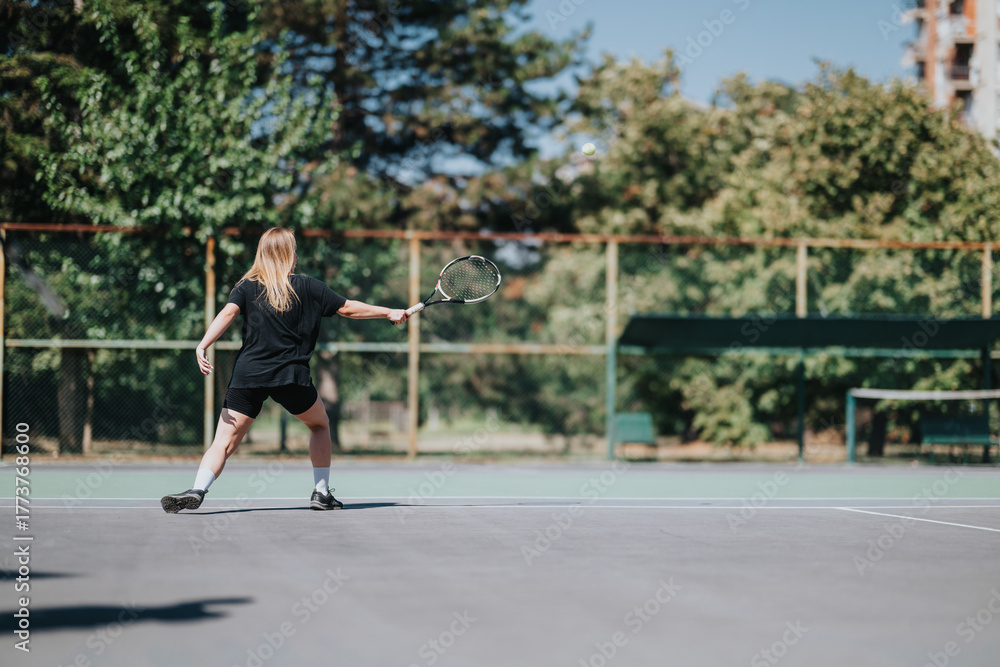 Fototapeta premium A woman in black athletic wear plays tennis on a bright outdoor court. She swings with focus as the ball flies by, framed by trees and fencing in a calm, sporty scene.