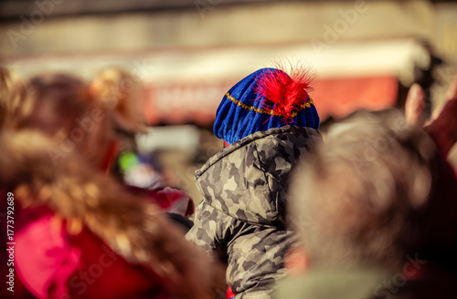 A young child wearing a blue hat sits on a parent’s shoulders, eagerly awaiting the arrival of Saint Nicholas during the festive Sinterklaas festivities in Dordrecht.