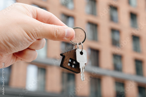man hand with metal key, keychain in shape of private house, modern brown brick multi-storey apartment building in background, purchase of real estate, real estate services