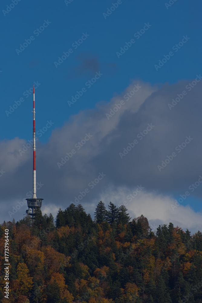 Fototapeta premium A tall red and white communications tower stands on a forested hilltop under a partly cloudy blue sky during the daytime.