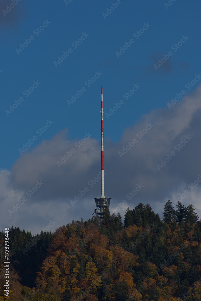 Fototapeta premium A tall red and white communications tower stands on a forested hilltop under a partly cloudy blue sky during the daytime.