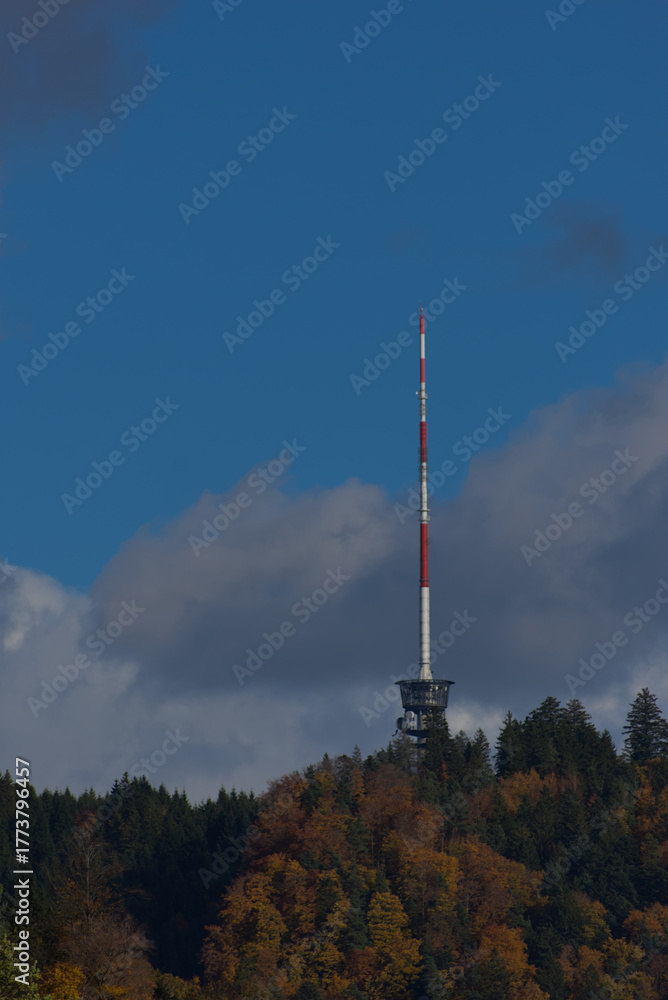Obraz premium A tall red and white communications tower stands on a forested hilltop under a partly cloudy blue sky during the daytime.