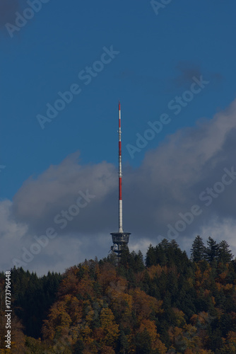 A tall red and white communications tower stands on a forested hilltop under a partly cloudy blue sky during the daytime.