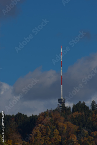 A tall red and white communications tower stands on a forested hilltop under a partly cloudy blue sky during the daytime.