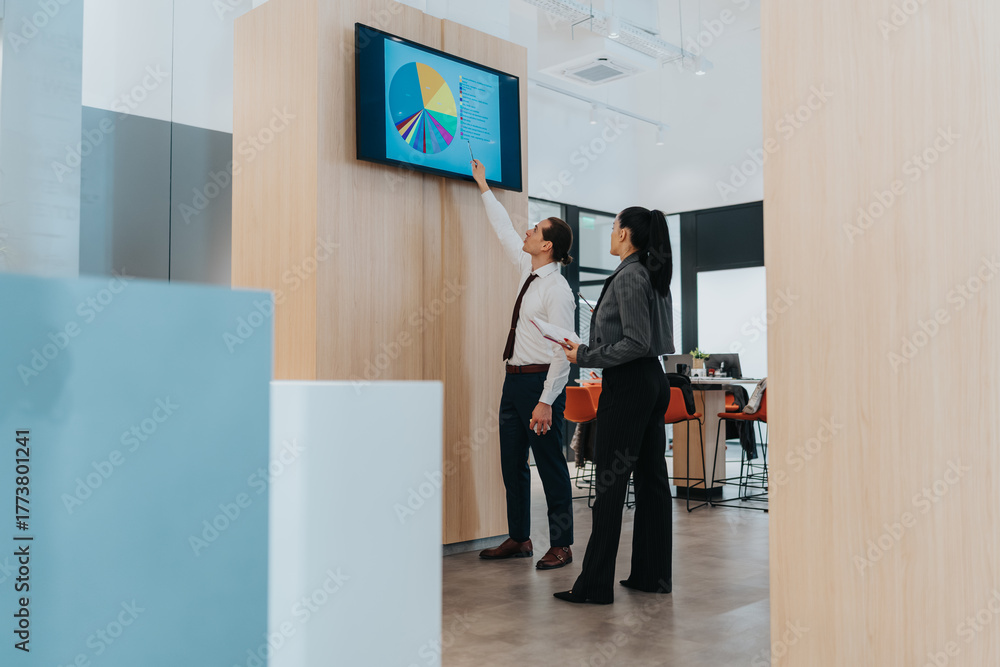 Fototapeta premium Two colleagues review a colorful dashboard on a wall screen inside a bright, modern office. They discuss data, pie charts, and strategy, highlighting teamwork, diversity, and business collaboration.