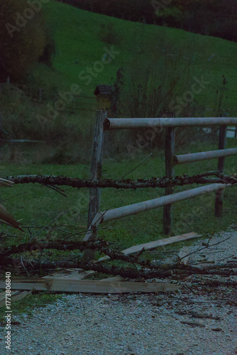 A wooden split-rail fence with broken and moss-covered sections borders a gravel path next to a grassy area and dark trees in the evening