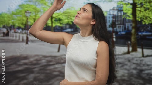 Hispanic woman in white sleeveless top shielding arm over eyes under bright sunlight on street; frustration.