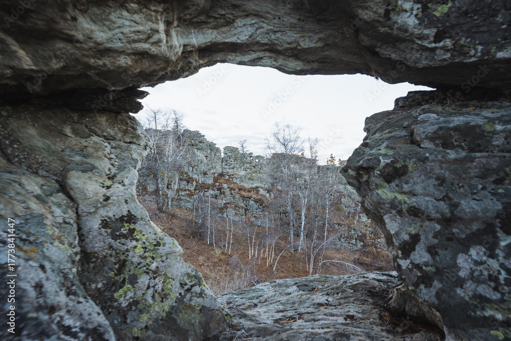 Fototapeta premium Natural rock window revealing forest basin below and distant ridge, textured arch with lichen framing panoramic vista, soft cool light and layered composition evoke calm remote landscape