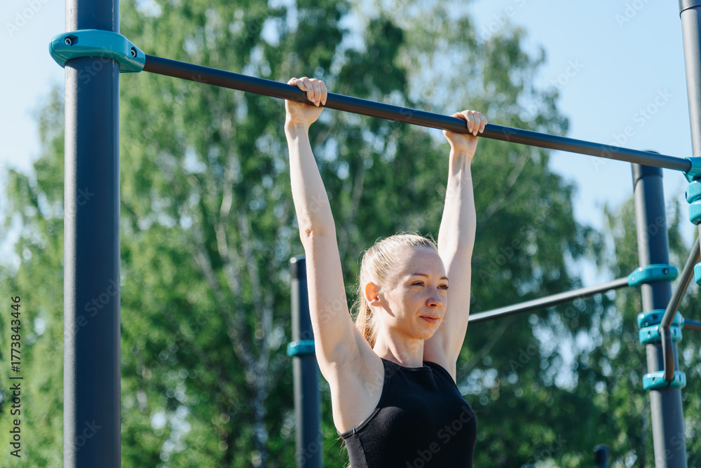 Fototapeta premium young sportive caucasian woman doing exercises on horizontal bar outdoors in summer, beautiful redhead girl, black sportswear, healthy lifestyle concept