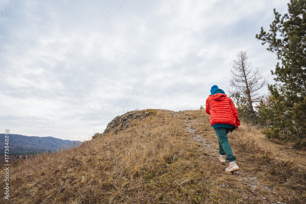 Fototapeta premium Young boy climbs rugged slope, Young child wearing colorful clothing moves across rugged terrain, Children in vibrant attire ascend challenging hill under overcast sky with determination