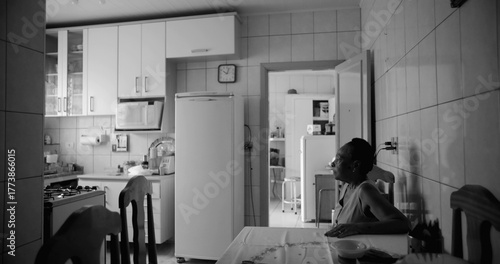 Fotomural Elderly woman sitting at a dining table in a tiled kitchen, gazing thoughtfully towards the light, in a serene and reflective moment, black and white