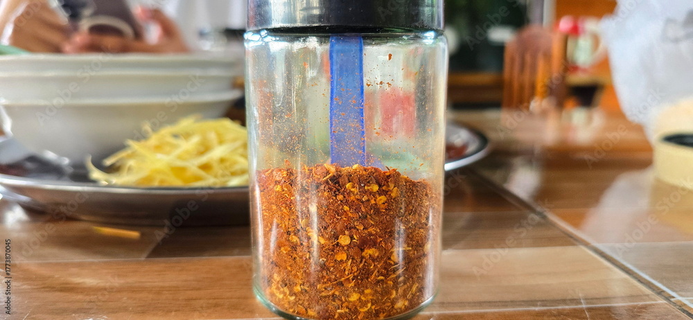 Obraz premium Chili powder is packaged in a clear bottle with a black cap and placed on the dining table. Crushed red pepper is a spicy seasoning. Dried chilies are finely ground to be used spicy seasoning in food.