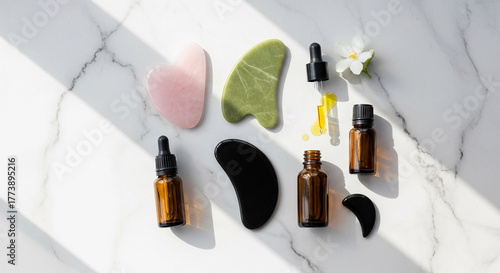 Gua sha tools and essential oils arranged on a marble surface. Includes pink, green, and black stone tools, along with amber dropper bottles for self-care and wellness routines.