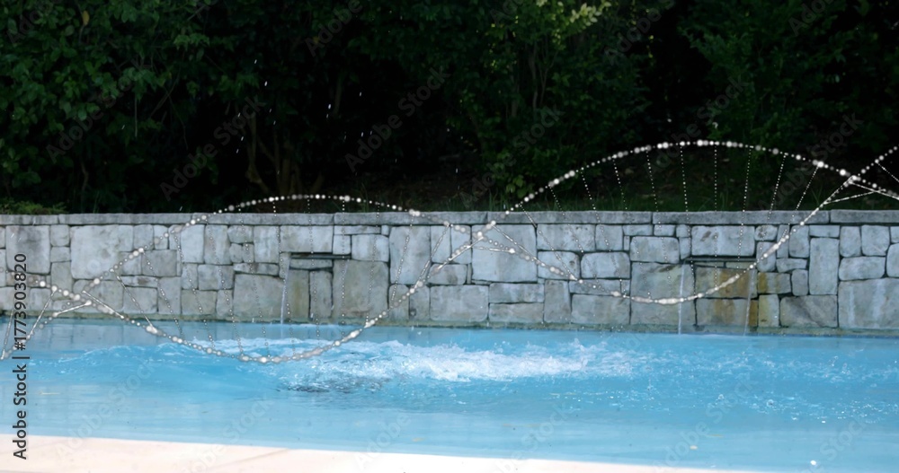 Fototapeta premium Arcing water jets splashing blue water at backyard pool, with stone wall fountains, concrete deck