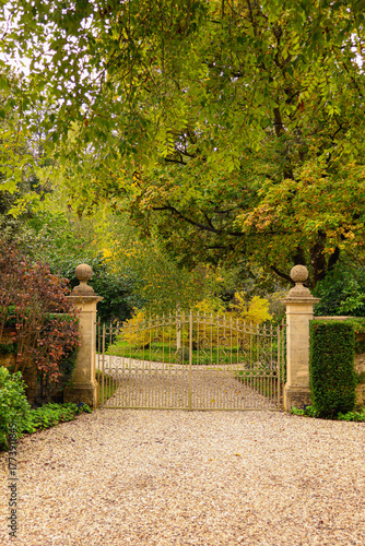 Estate entrance with closed golden ornate gate, gravel driveway, and lush autumn garden. Cotswolds countryside, England