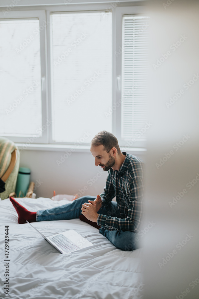Obraz premium A man wearing a plaid shirt and red socks sits on a bed working on a laptop in a casual home environment. He displays focus and engagement during this remote working moment.