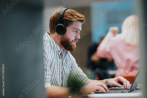 Obraz na płótnie A bearded man wearing large over-ear headphones concentrates on his laptop at a modern desk