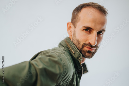 Schilderij op canvas Close-up captured image of a man with a confident demeanor, wearing a green jacket, looking directly at the viewer in a bright and minimal setting, portraying self-assurance and approachability