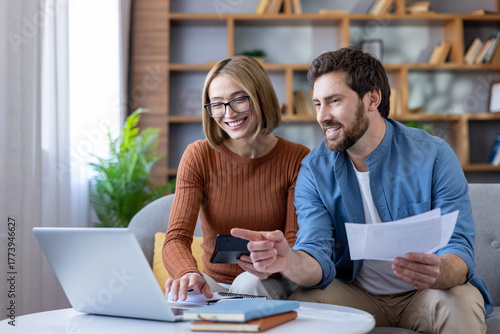 Smiling couple sitting on a sofa, planning their household expenses and investments, calculating figures with a laptop and calculator, organizing bills and paperwork together in their living room