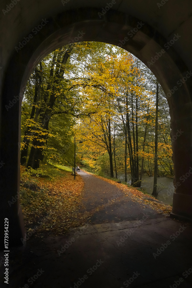 Fototapeta premium Autumn park path through archway. Scenic walking trail through tunnel to park with yellow fall leaves.